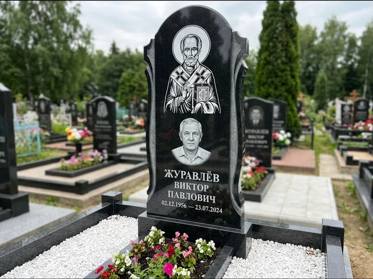 Памятник с гравировкой иконы: стела с образом в полный рост на обороте — гравировка образа Матроны Московской, низкие кусты спиреи в цвету, работа «Лебедева» в Хабаровске